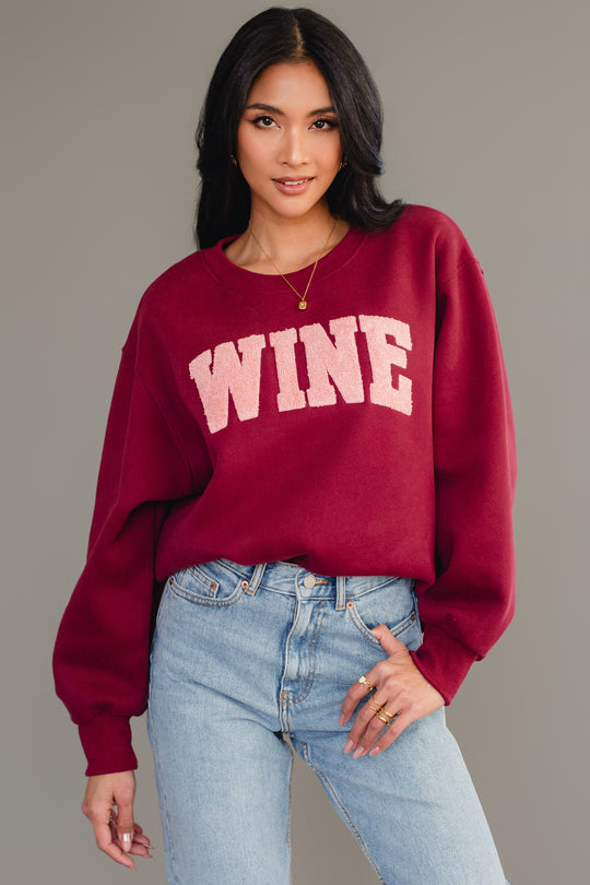 A woman with long dark hair models the Panache Apparel Collection Burgundy Wine Sweatshirt with chenille "WINE" lettering, paired with light blue jeans, against a plain gray background.