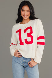 A woman models the Cream & Red 13 Sweater from Panache Apparel Collection, featuring bold red number 13 and sleeve stripes, styled with light blue jeans as she smiles at the camera against a plain gray background.