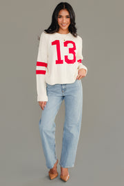 A woman wearing the Panache Apparel Collection Cream & Red 13 Sweater, light blue jeans, and tan heels stands smiling against a plain gray background.