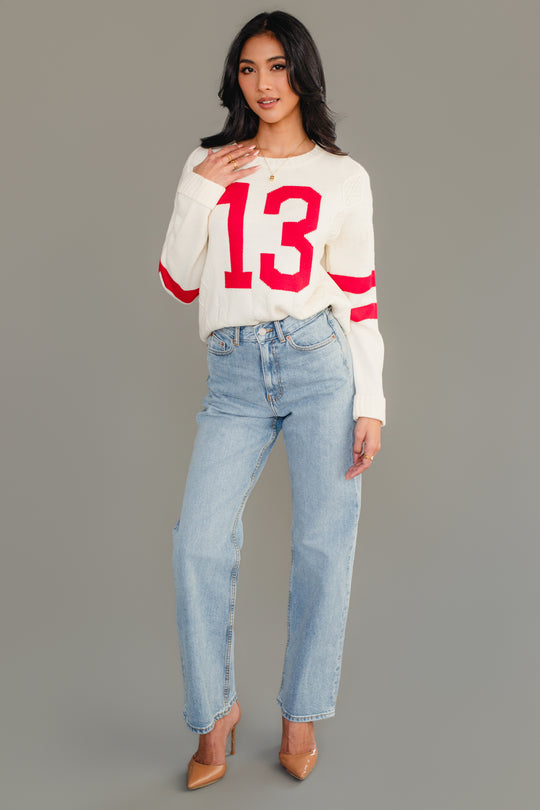 A woman with long black hair stands against a gray background, gently touching her Cream & Red 13 Sweater from Panache Apparel Collection. She pairs the sweater with light blue jeans and tan heels.