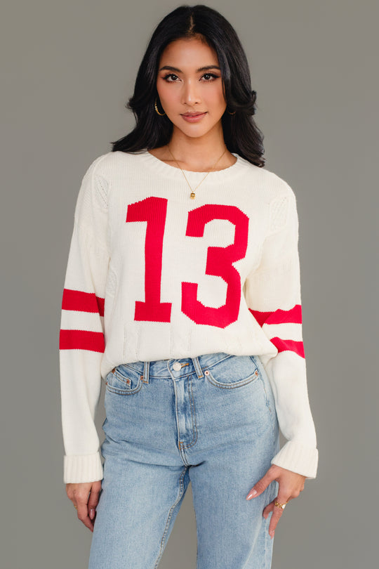 A woman models the Panache Apparel Collection's Cream & Red 13 Sweater, featuring bold red sleeve stripes and the number 13 on the front, paired with light blue jeans against a plain gray background.
