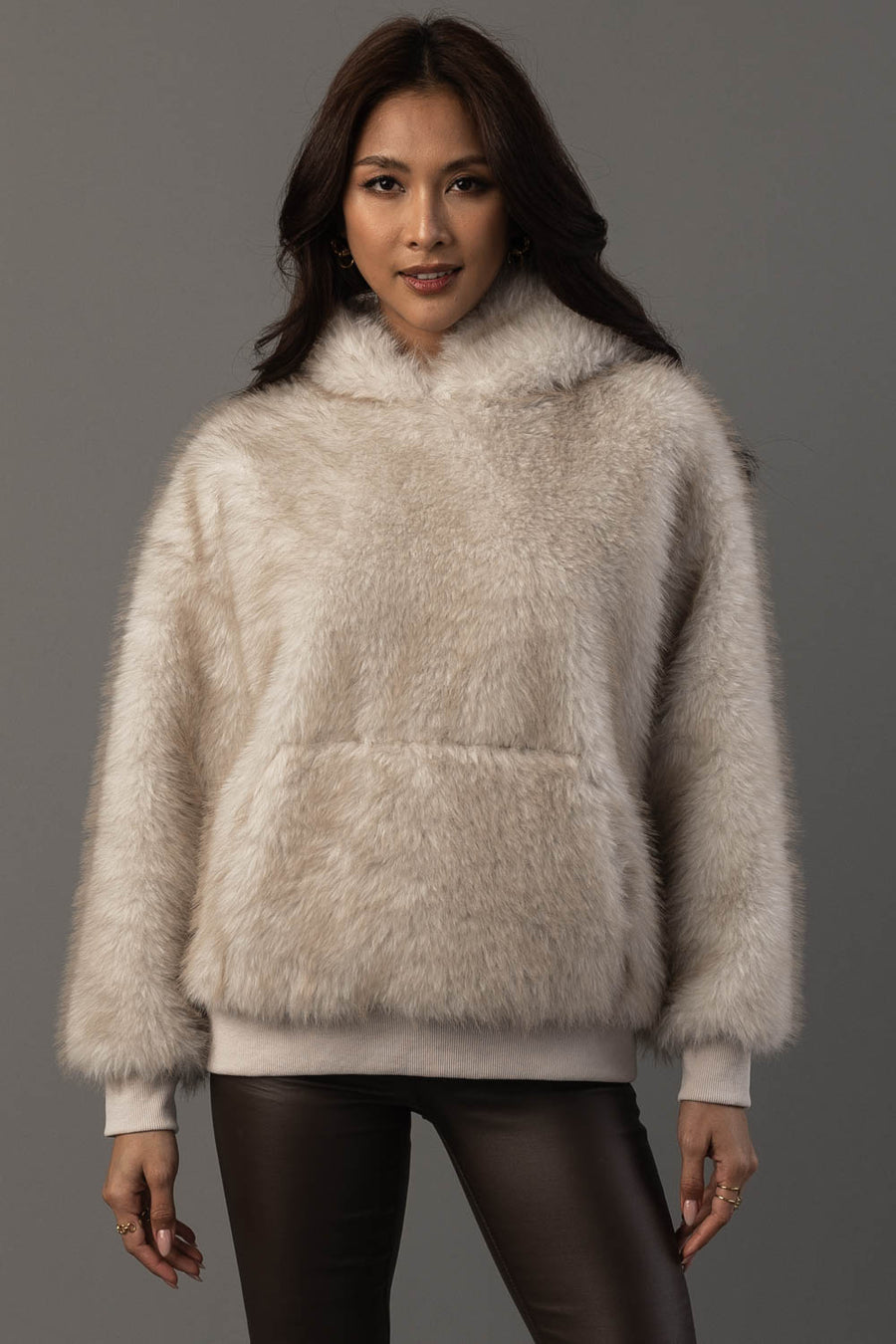 A woman stands against a gray background, facing forward with her hands relaxed by her sides, wearing the Panache Apparel Collection Cream Faux Fur Hooded Sweatshirt and dark pants.