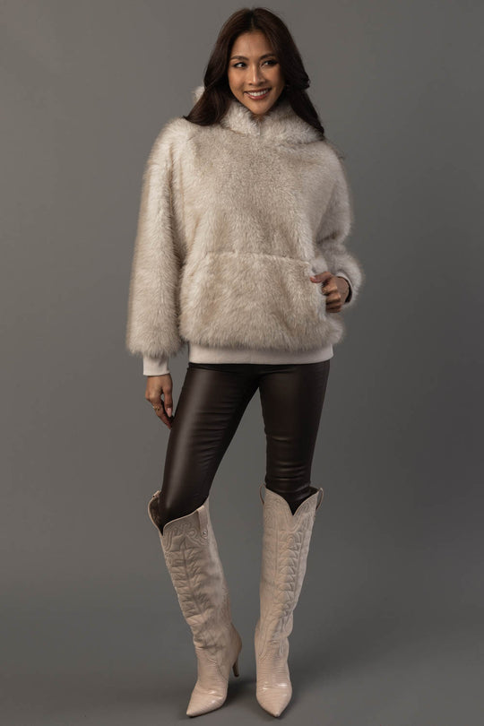 A woman models the Panache Apparel Collection Cream Faux Fur Hooded Sweatshirt, paired with black leather leggings and cream-colored cowboy boots, against a plain gray background.