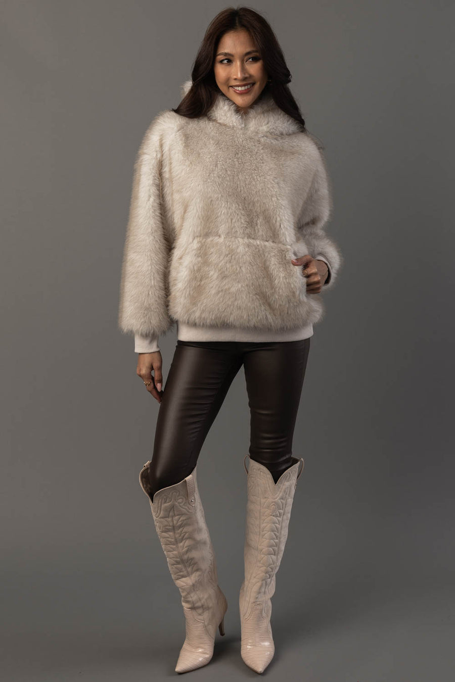 A woman models the Panache Apparel Collection Cream Faux Fur Hooded Sweatshirt, paired with black leather leggings and cream-colored cowboy boots, against a plain gray background.