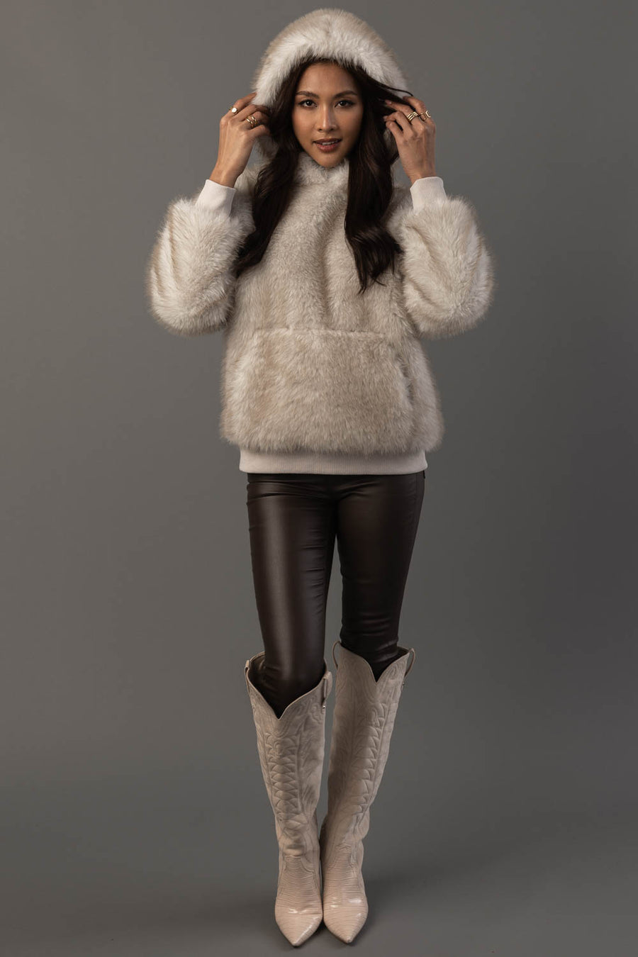 A woman stands against a gray background, wearing the Panache Apparel Collection Cream Faux Fur Hooded Sweatshirt, paired with black leather pants and cream-colored knee-high cowboy boots. She is holding the hood with both hands and smiling.