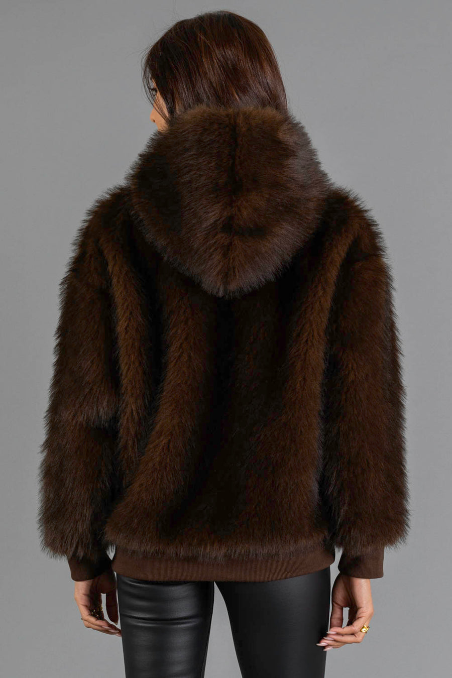 A person stands facing away, wearing the Panache Apparel Collection Dark Brown Faux Fur Hooded Sweatshirt and black leather pants, against a plain gray background.