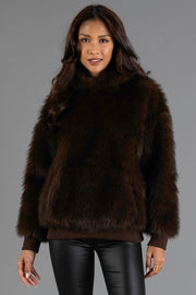A woman wears the Panache Apparel Collection Dark Brown Faux Fur Hooded Sweatshirt paired with black leather pants, standing against a plain gray background and smiling softly at the camera.