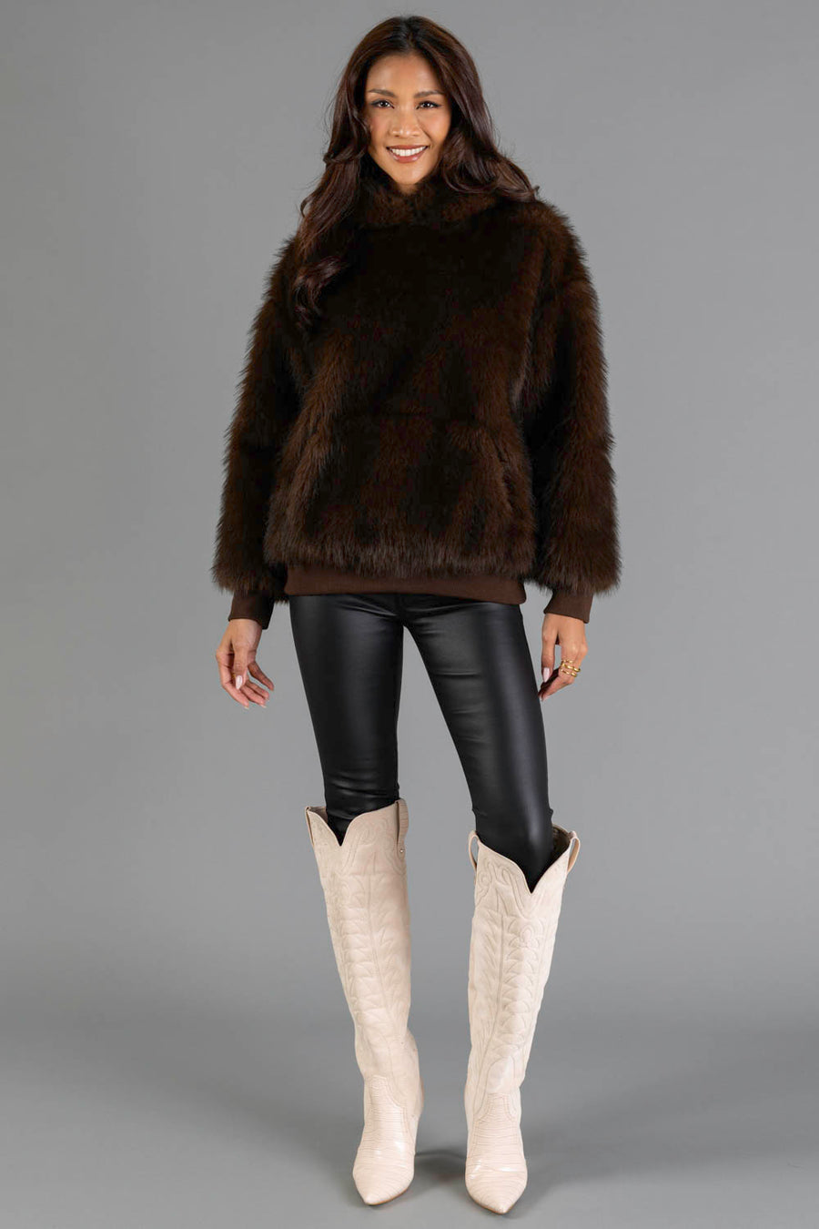 A woman stands smiling in a Panache Apparel Collection Dark Brown Faux Fur Hooded Sweatshirt, paired with black leather leggings and tall white cowboy boots, against a plain gray background.