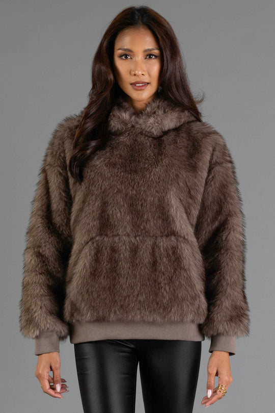 A woman with long dark hair models the Gray/Brown Faux Fur Hooded Sweatshirt from Panache Apparel Collection, paired with black leather pants and set against a plain gray background.