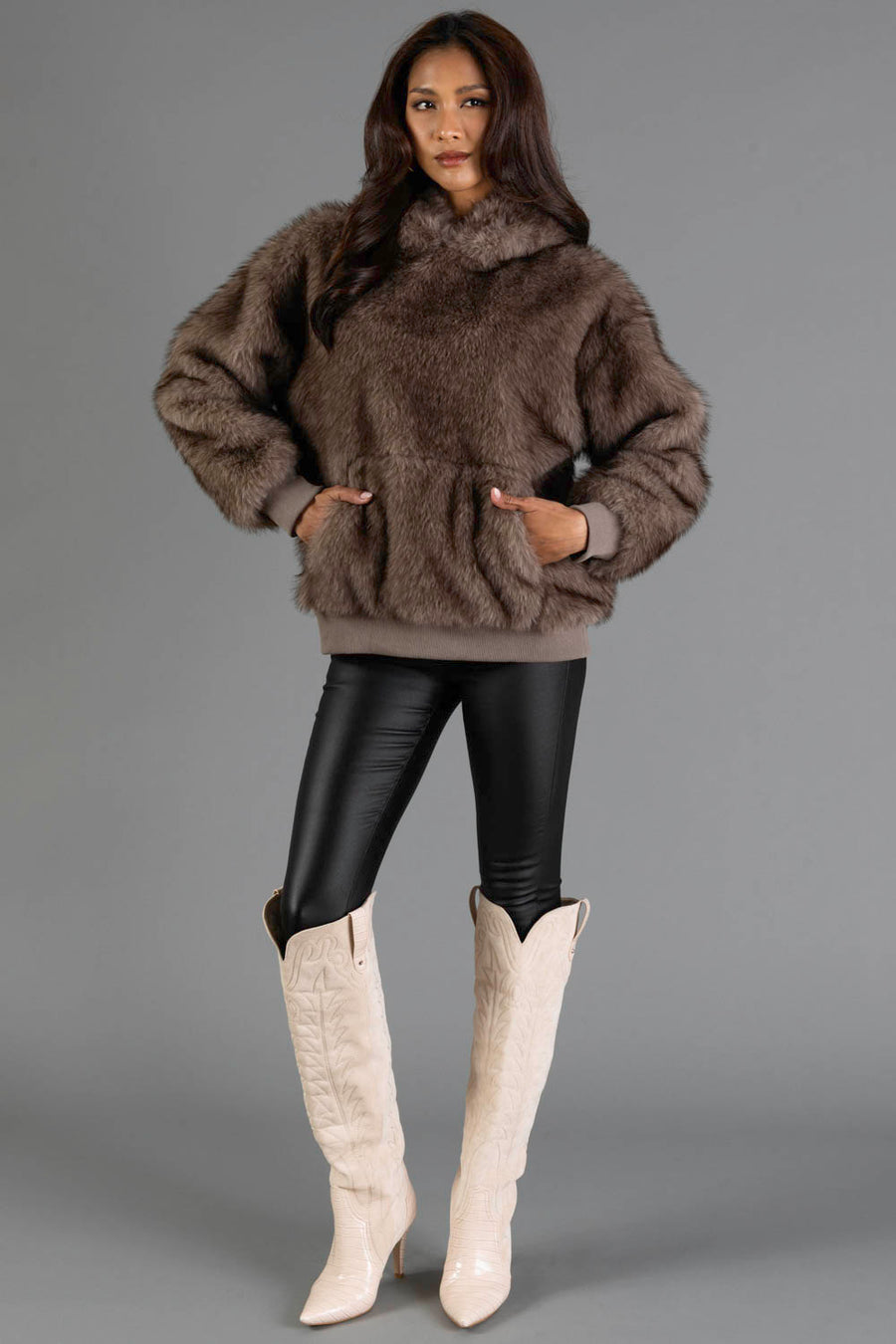 A woman wearing the Gray/Brown Faux Fur Hooded Sweatshirt by Panache Apparel Collection, paired with black leather leggings and cream cowboy boots, stands against a plain gray background, facing forward with her hands in the sweatshirt pockets.
