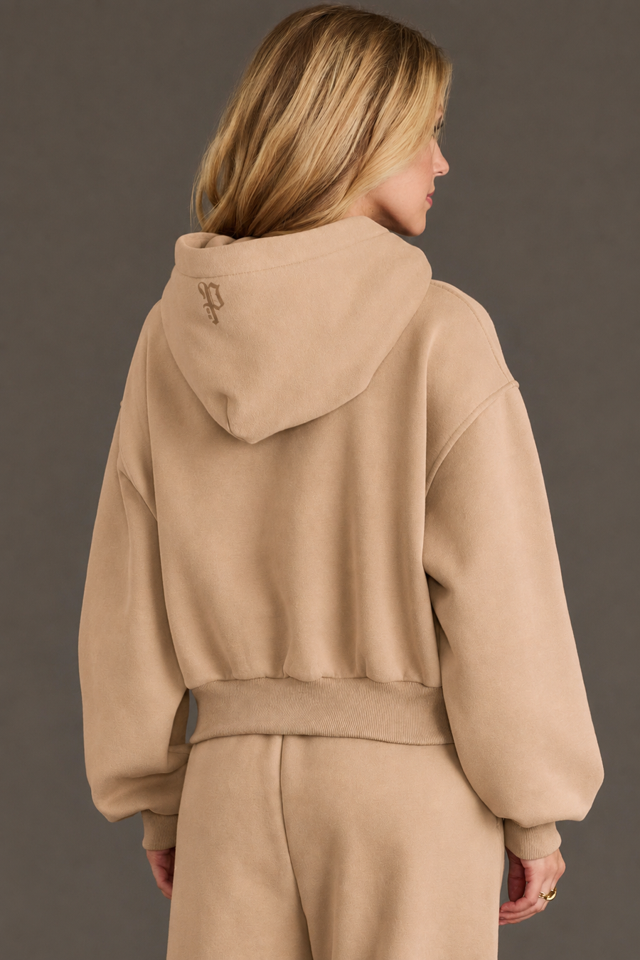 A woman with blonde hair is seen from behind wearing the Khaki Panache Cropped Hooded Sweatshirt and matching pants from the Panache Apparel Collection. The fleece sweatshirt includes a hood with a small, embroidered logo at the center. She stands against a plain, neutral background.
