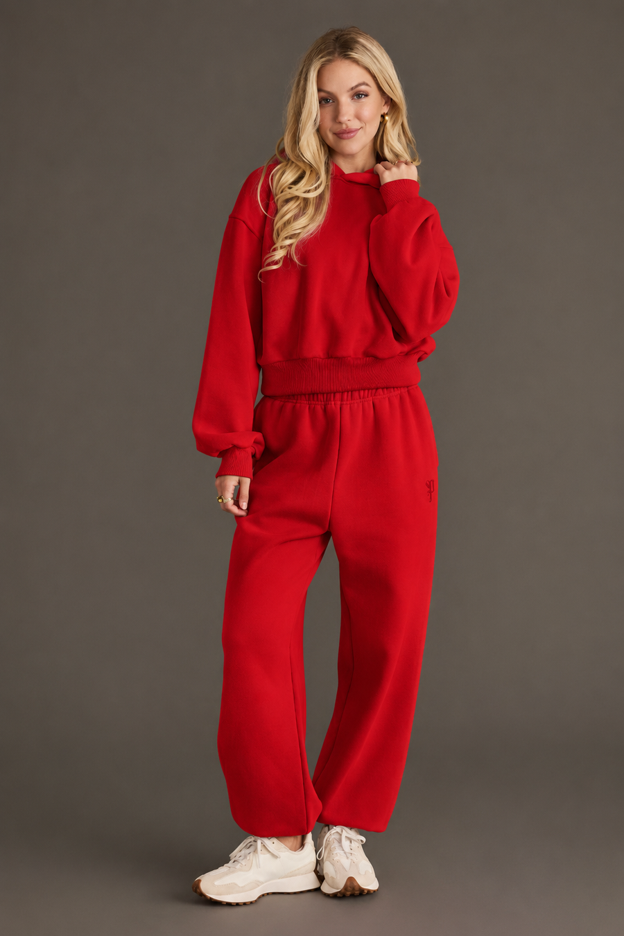 A woman with long blonde hair stands smiling with one hand touching her hair, wearing the Panache Apparel Collection’s Red Panache Cropped Hooded Sweatshirt and matching red sweatpants, paired with white sneakers against a plain background.