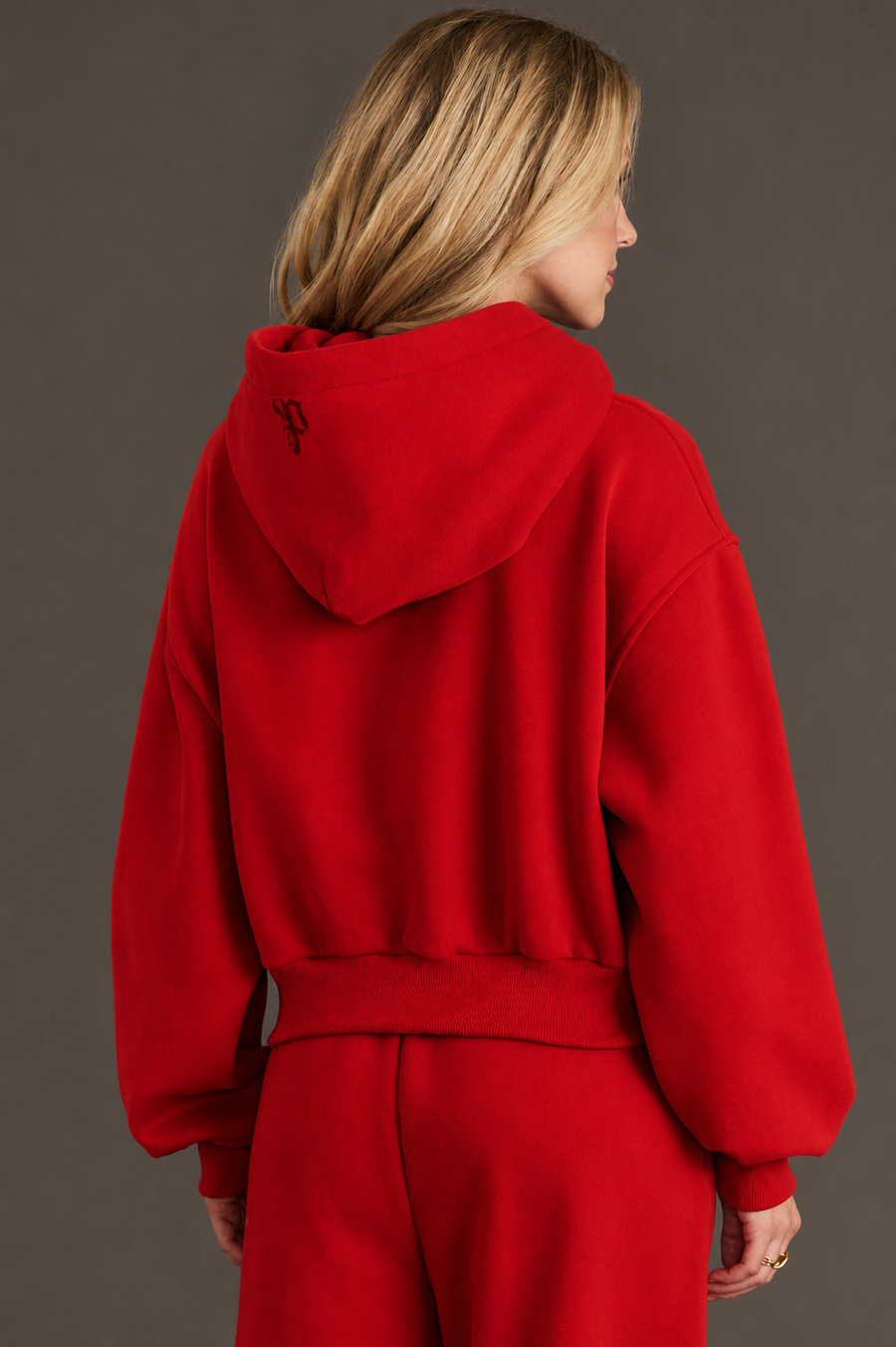 A person with blonde hair, facing away from the camera, is wearing a Red Panache Cropped Hooded Sweatshirt and matching pants from the Panache Apparel Collection. A small black logo is visible on the back of the hood. The background is plain and dark.
