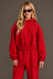 Wearing the Red Panache Cropped Hooded Sweatshirt from Panache Apparel Collection with matching red sweatpants and black sunglasses, a person stands against a plain, dark background, smiling slightly with their hands relaxed by their sides.