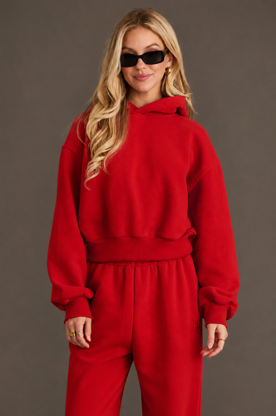 Wearing the Red Panache Cropped Hooded Sweatshirt from Panache Apparel Collection with matching red sweatpants and black sunglasses, a person stands against a plain, dark background, smiling slightly with their hands relaxed by their sides.