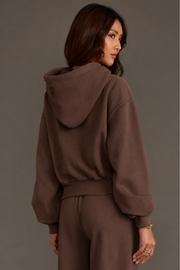A woman with long brown hair is wearing the Panache Apparel Collection’s Brown Panache Cropped Hooded Sweatshirt, featuring a cozy fleece interior and loose fit, paired with matching pants as she faces away and looks over her shoulder against a plain gray background.