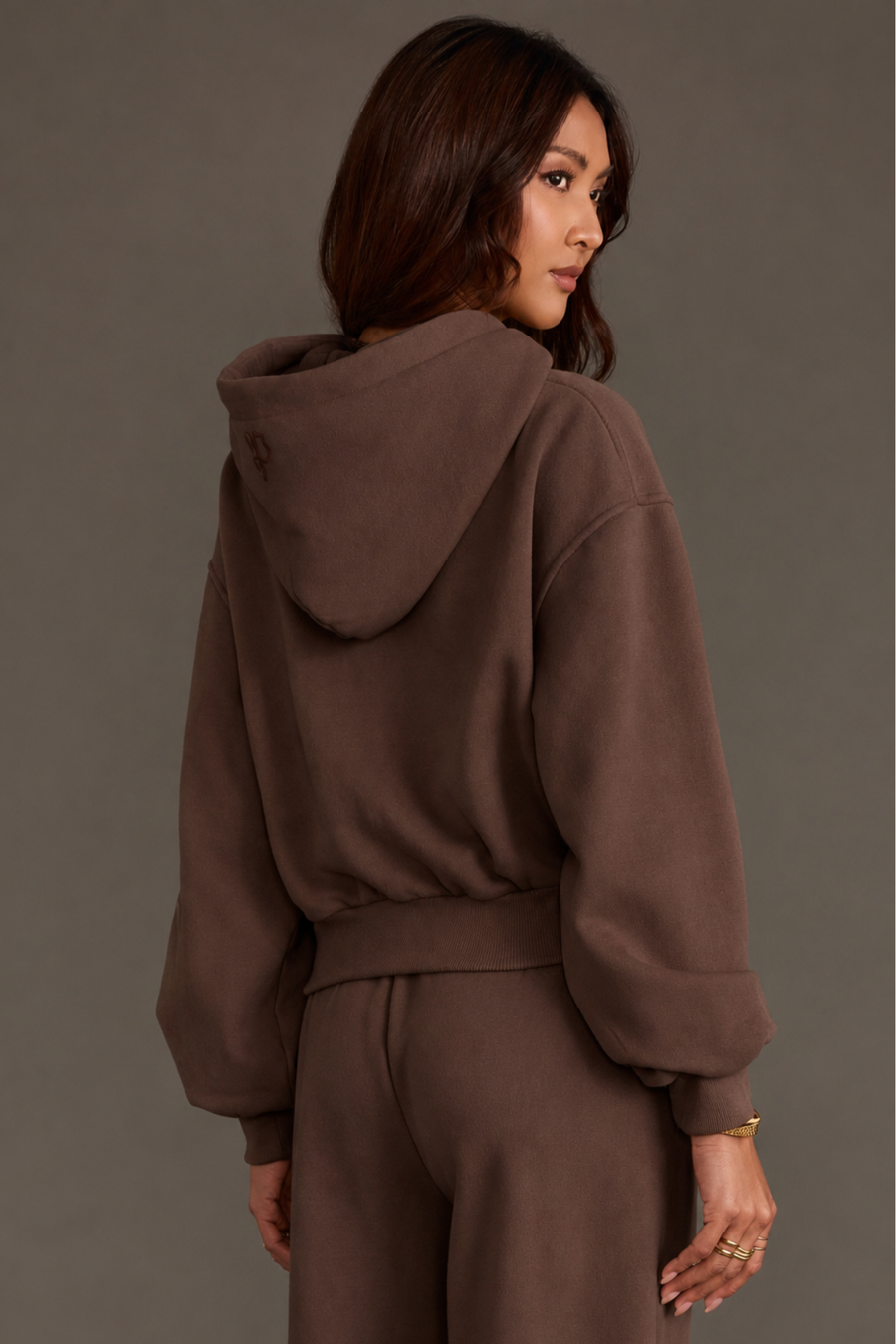 A woman with long brown hair is wearing the Panache Apparel Collection’s Brown Panache Cropped Hooded Sweatshirt, featuring a cozy fleece interior and loose fit, paired with matching pants as she faces away and looks over her shoulder against a plain gray background.