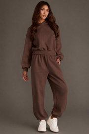 A woman stands against a plain gray background wearing the Panache Apparel Collection’s Brown Panache Cropped Hooded Sweatshirt styled with matching sweatpants, both featuring a relaxed fit and subtle brown flair, completed with white sneakers.