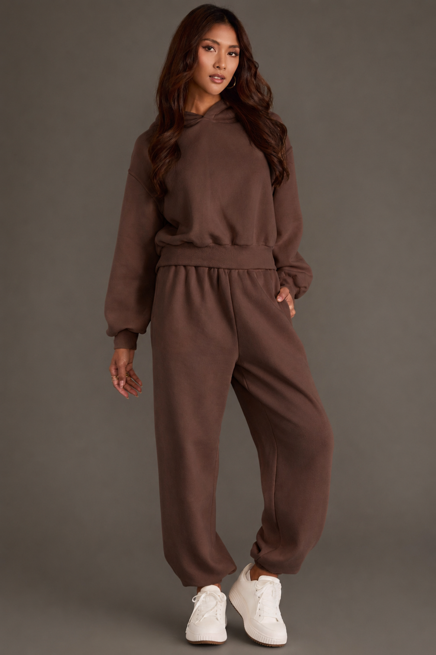 A woman stands against a plain gray background wearing the Panache Apparel Collection’s Brown Panache Cropped Hooded Sweatshirt styled with matching sweatpants, both featuring a relaxed fit and subtle brown flair, completed with white sneakers.
