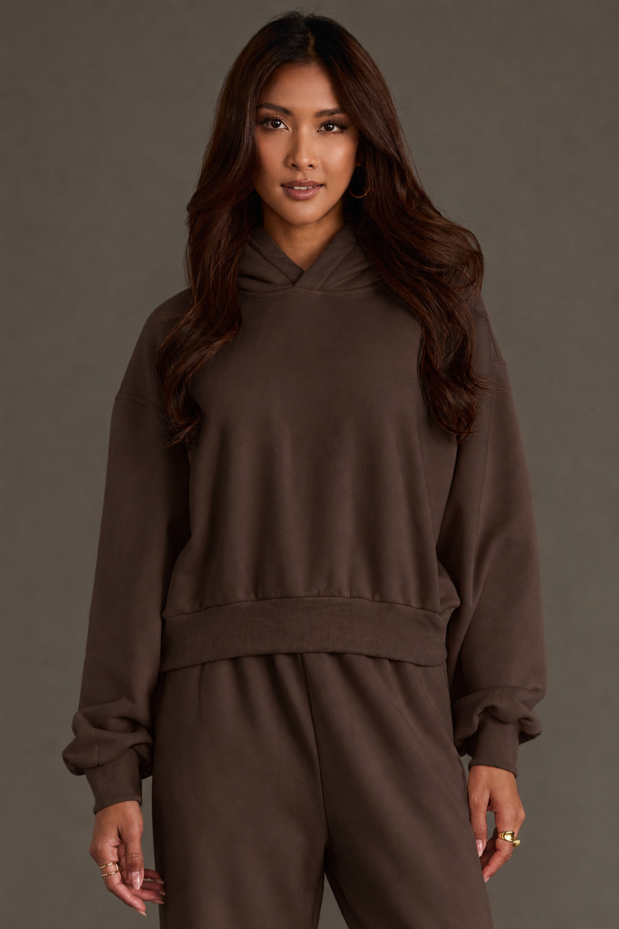 A woman with long, wavy brown hair models the Panache Apparel Collection’s Brown Panache Cropped Hooded Sweatshirt, showcasing its cozy fleece interior and matching pants while standing against a plain gray background and gazing confidently at the camera.