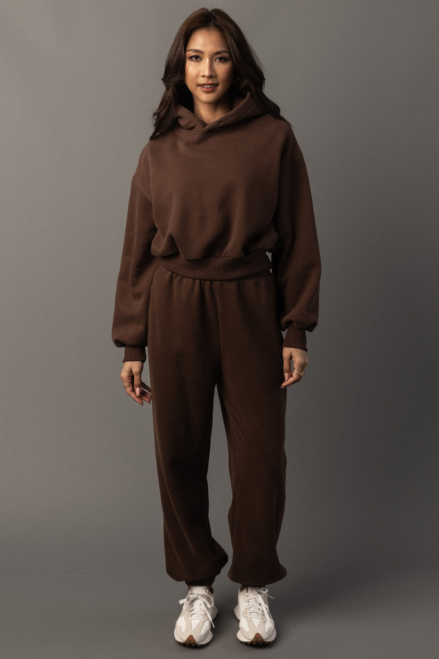 A person stands against a gray background wearing the Brown Panache Cropped Hooded Sweatshirt from the Panache Apparel Collection, along with matching sweatpants and white sneakers, smiling slightly with their hands relaxed by their sides.