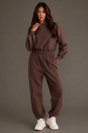 A woman models the Brown Panache Cropped Hooded Sweatshirt from the Panache Apparel Collection, pairing the long-sleeved top with matching pants. With long hair and white sneakers, she poses against a plain gray background with one hand near her face.