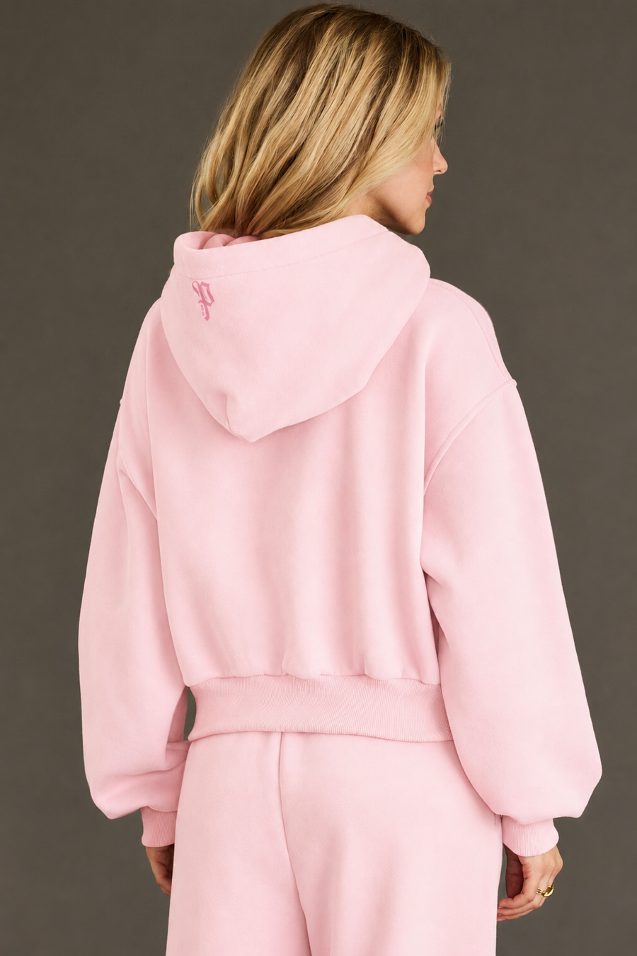 A person with blonde hair wears the Light Pink Panache Cropped Hooded Sweatshirt from Panache Apparel Collection, paired with matching sweatpants, facing away from the camera against a plain, dark background.