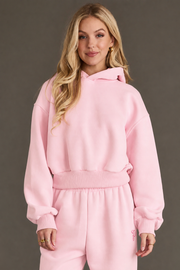 A person with long blonde hair wears a Light Pink Panache Cropped Hooded Sweatshirt and matching sweatpants from the Panache Apparel Collection, standing against a plain dark background and smiling slightly at the camera.