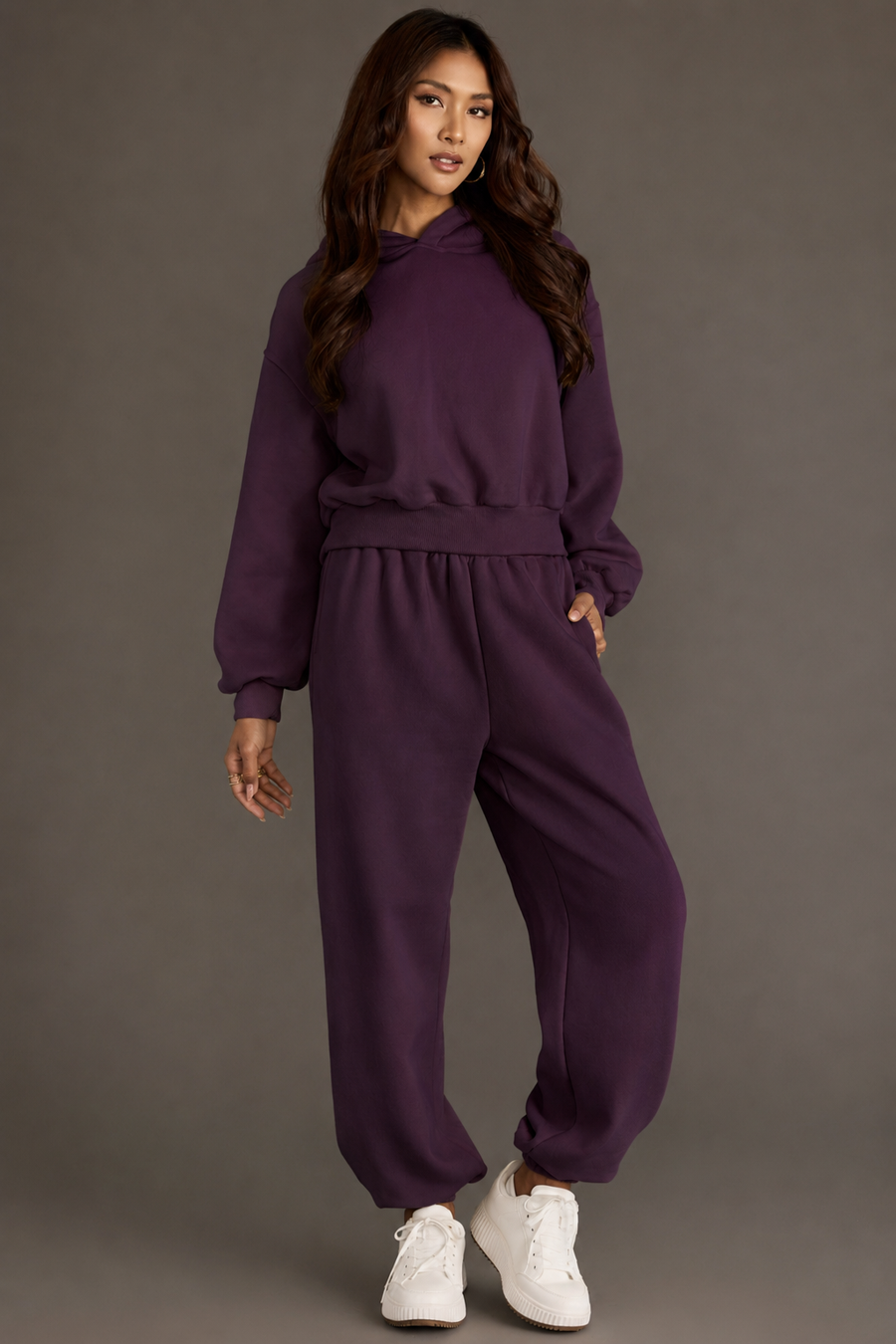 Dressed in the Plum Panache Cropped Hooded Sweatshirt from the Panache Apparel Collection and matching relaxed fit sweatpants, a woman stands against a gray background, posing with one hand in her pocket and sporting white sneakers. The soft fleece fabric elevates her effortlessly stylish look with added comfort.