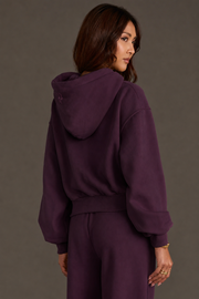 A woman with long brown hair models the Plum Panache Cropped Hooded Sweatshirt from the Panache Apparel Collection, featuring a relaxed fit in soft fleece and paired with matching pants, as she looks over her shoulder against a plain background.