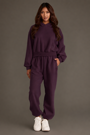 A person stands facing forward, dressed in the Plum Panache Cropped Hooded Sweatshirt from Panache Apparel Collection—a relaxed fit, dark purple fleece hoodie—paired with matching sweatpants and white sneakers, set against a plain, neutral background.
