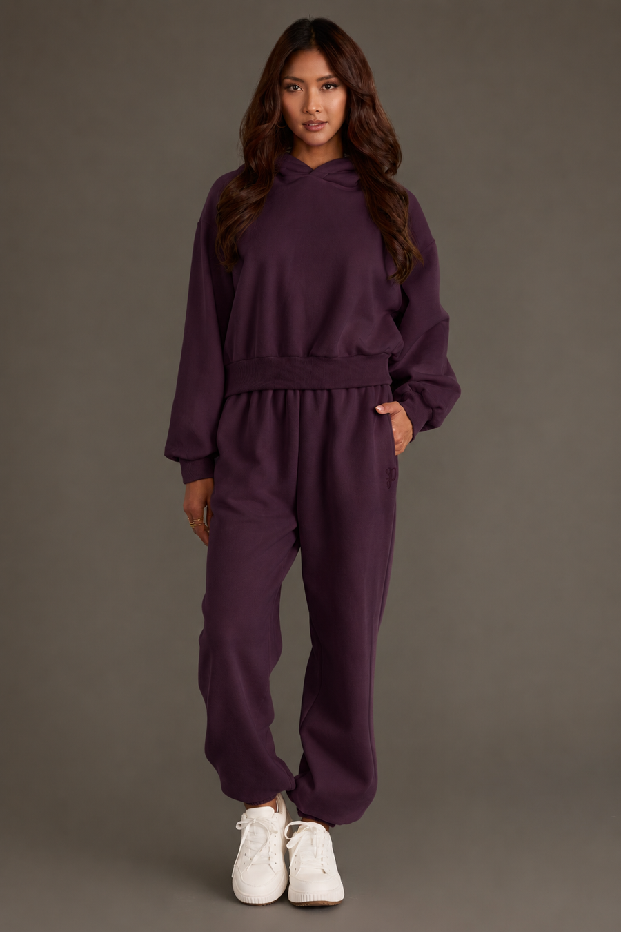 A person stands facing forward, dressed in the Plum Panache Cropped Hooded Sweatshirt from Panache Apparel Collection—a relaxed fit, dark purple fleece hoodie—paired with matching sweatpants and white sneakers, set against a plain, neutral background.
