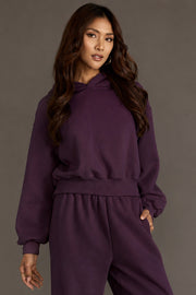 A person with long, wavy brown hair wears the Plum Panache Cropped Hooded Sweatshirt from Panache Apparel Collection, paired with matching dark purple fleece sweatpants, standing against a plain gray background with one hand resting on their hip.