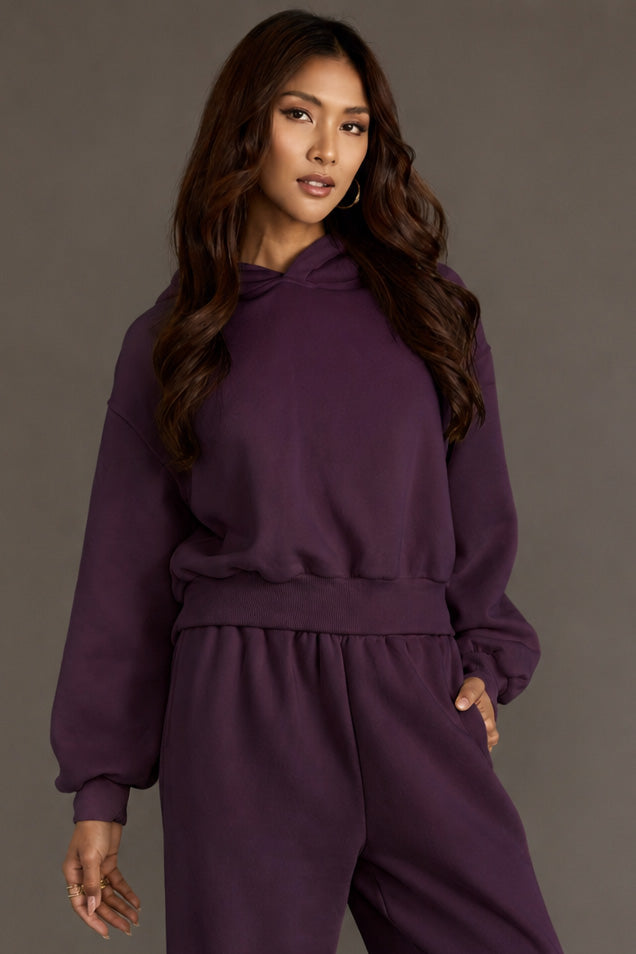 A person with long, wavy brown hair wears the Plum Panache Cropped Hooded Sweatshirt from Panache Apparel Collection, paired with matching dark purple fleece sweatpants, standing against a plain gray background with one hand resting on their hip.