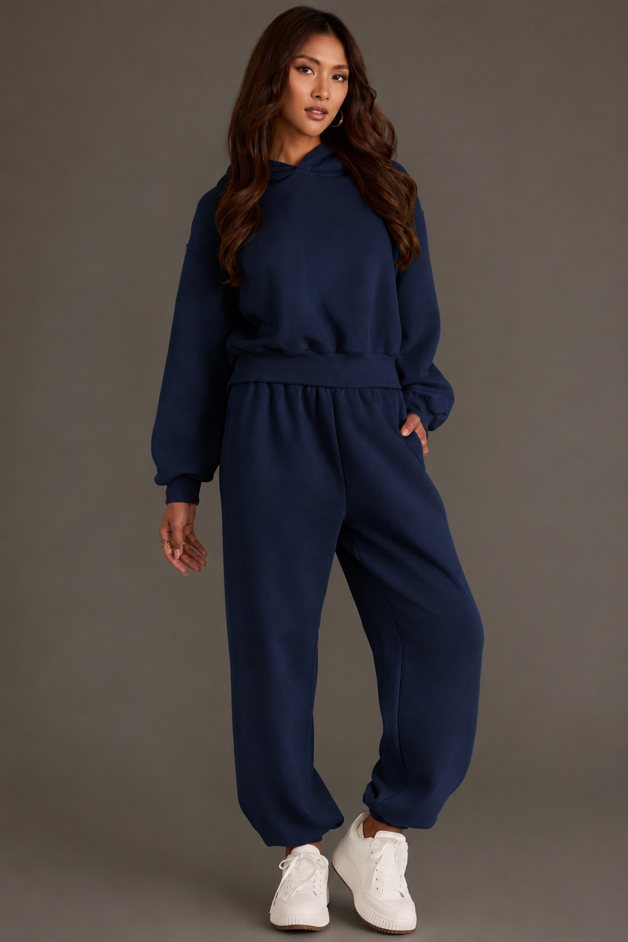 Wearing the Dark Navy Panache Cropped Hooded Sweatshirt from the Panache Apparel Collection and matching jogger pants, a woman poses in white sneakers against a plain gray background.