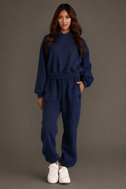 A woman stands facing forward, wearing a Dark Navy Panache Cropped Hooded Sweatshirt from the Panache Apparel Collection, paired with matching jogger pants and white sneakers, against a plain dark background.
