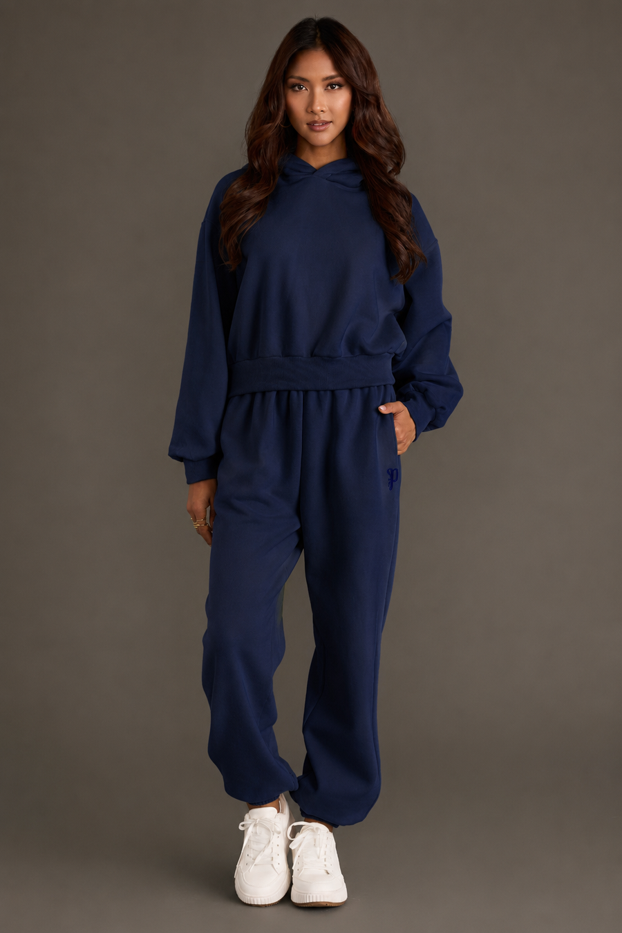 A woman stands facing forward, wearing a Dark Navy Panache Cropped Hooded Sweatshirt from the Panache Apparel Collection, paired with matching jogger pants and white sneakers, against a plain dark background.