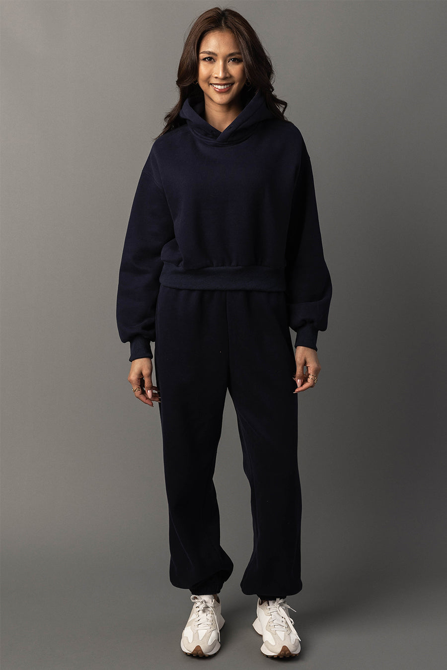A smiling person stands against a gray background, wearing the Panache Apparel Collection’s Dark Navy Panache Cropped Hooded Sweatshirt, matching sweatpants, and white sneakers.