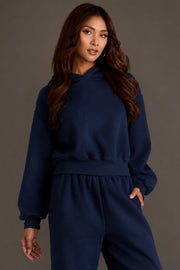 A woman wearing the Panache Apparel Collection’s Dark Navy Panache Cropped Hooded Sweatshirt stands against a plain gray background, looking toward the camera with a neutral expression.