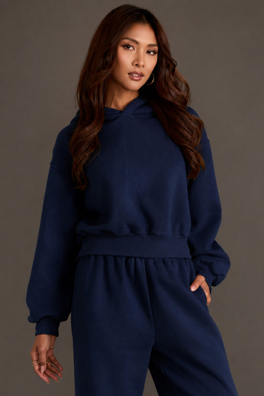 A woman wearing the Panache Apparel Collection’s Dark Navy Panache Cropped Hooded Sweatshirt stands against a plain gray background, looking toward the camera with a neutral expression.