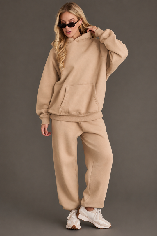 A woman wearing the Khaki Panache Hooded Sweatshirt from the Panache Apparel Collection with matching sweatpants, white sneakers, black sunglasses, and hoop earrings stands against a plain gray background.