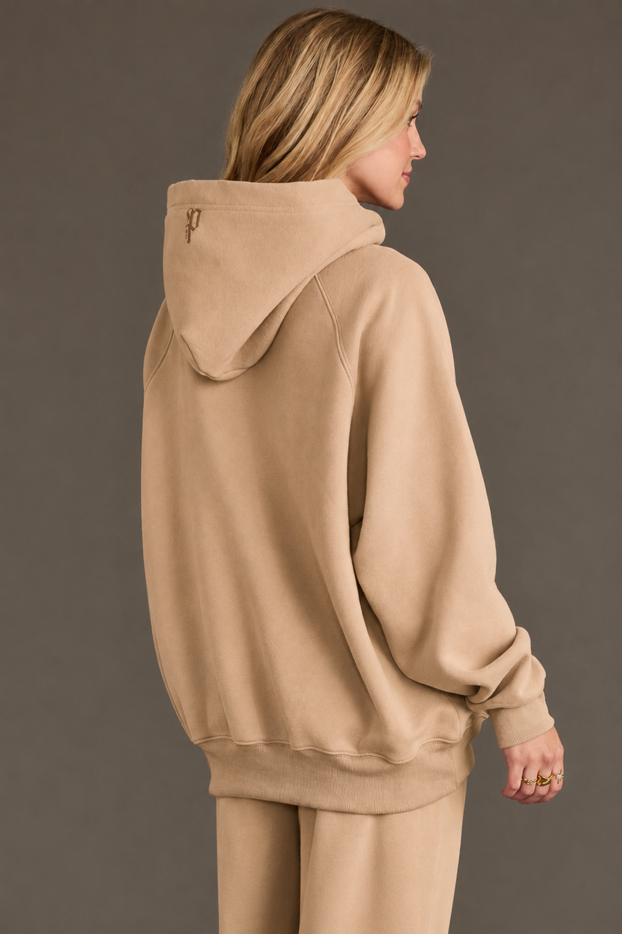 A woman with blonde hair faces away, wearing the Panache Apparel Collection’s Khaki Panache Hooded Sweatshirt in a soft tan shade with the hood up and matching pants. The background is plain and neutral.