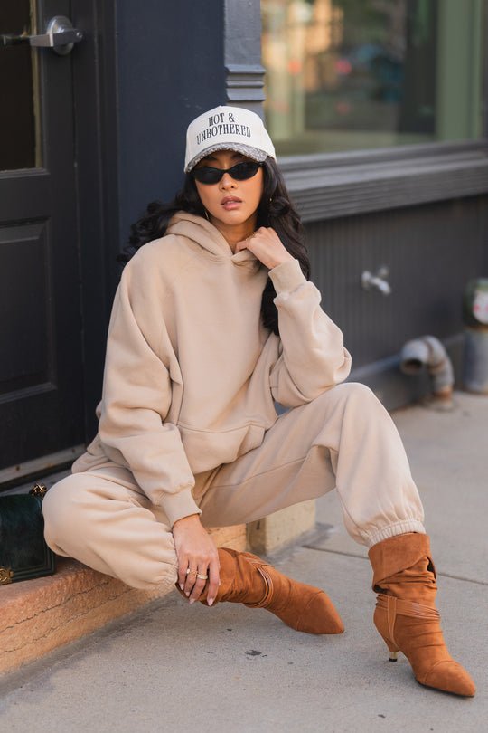 A person wearing a Khaki Panache Hooded Sweatshirt and matching sweatpants, a baseball cap that reads HOT & UNBOTHERED, sunglasses, and brown pointed-toe heeled boots, sitting on a step.