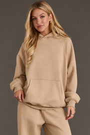 A woman with long blonde hair wears the Khaki Panache Hooded Sweatshirt from Panache Apparel Collection and matching sweatpants, standing against a plain gray background with one hand in her pocket and a slight smile on her face.