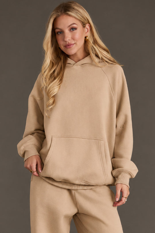 A woman with long blonde hair wears the Khaki Panache Hooded Sweatshirt from Panache Apparel Collection and matching sweatpants, standing against a plain gray background with one hand in her pocket and a slight smile on her face.