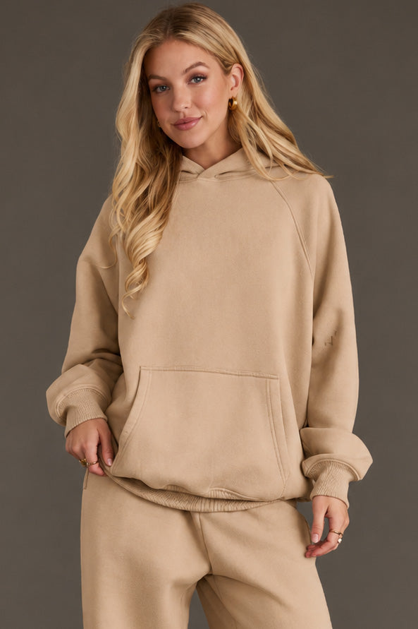 A woman with long blonde hair wears the Khaki Panache Hooded Sweatshirt from Panache Apparel Collection and matching sweatpants, standing against a plain gray background with one hand in her pocket and a slight smile on her face.