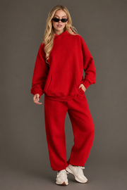 A woman dressed in the Panache Apparel Collection Red Panache Hooded Sweatshirt, paired with matching sweatpants, white sneakers, and sunglasses stands confidently against a gray background.