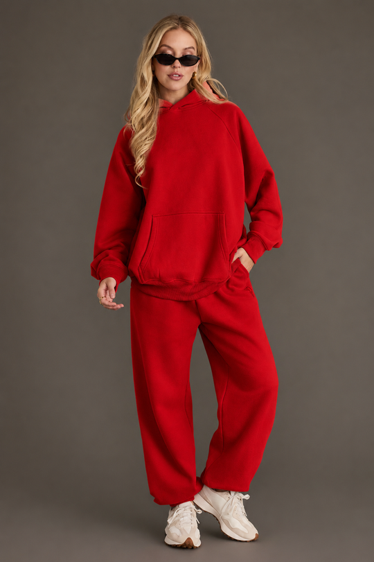 A woman dressed in the Panache Apparel Collection Red Panache Hooded Sweatshirt, paired with matching sweatpants, white sneakers, and sunglasses stands confidently against a gray background.
