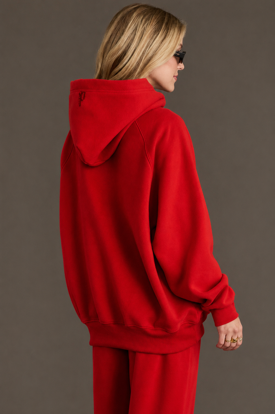 A person with blonde hair, wearing black sunglasses and the hood up on a Panache Apparel Collection Red Panache Hooded Sweatshirt, along with matching red pants, stands facing sideways against a plain background.
