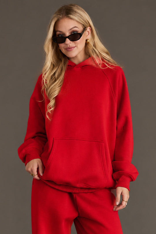 A woman with long blonde hair, wearing black sunglasses and the Red Panache Hooded Sweatshirt from Panache Apparel Collection—featuring a bright red color and soft fleece interior—paired with matching red sweatpants, stands against a plain gray background with her hands in the hoodie pocket.